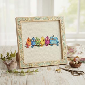 Finished Rainbow Birds Cross Stitch Pattern displayed in a hoop shows seven bright birds perched on a wire, each stitched in a different rainbow shade. Their playful personalities include singing, winking, and dozing off, making the design full of charm. The vibrant palette features oranges, yellows, blues, greens, and purples, balanced with backstitch outlines for clarity. This counted embroidery PDF pattern is perfect for stitchers who love cheerful and whimsical art, creating a handmade