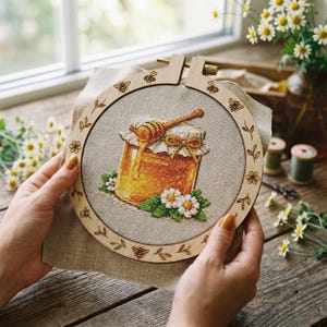 May include: Cross-stitch embroidery of a honey jar with a wooden honey dipper, surrounded by white flowers and green leaves. The embroidery is set in a wooden hoop with bee and floral designs. The background is a neutral linen fabric.