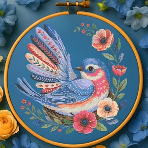 May include: A cross-stitch embroidery of a bluebird with colorful wings, nestled in a woven nest, surrounded by pink and peach flowers and green leaves. The embroidery is set in a wooden hoop against a blue background.