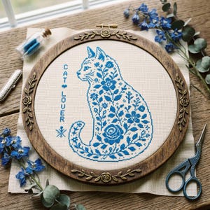 May include: A cross-stitch embroidery hoop featuring a blue floral cat design on cream fabric. The wooden hoop has decorative metal accents. The text "CAT LOVER" is stitched vertically. Blue scissors and flowers are nearby.
