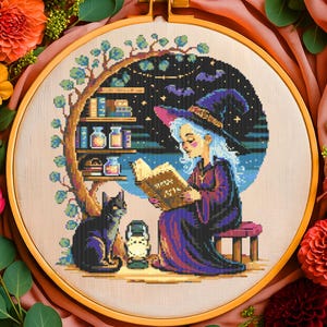 以下が含まれることがあります： 木製のフレームに入ったクロスステッチ刺繍。本を読む魔女、黒猫、ランタンが描かれています。魔女は紫色のドレスと帽子を着用。左側には本棚と木があり、夜空にはコウモリが飛んでいます。