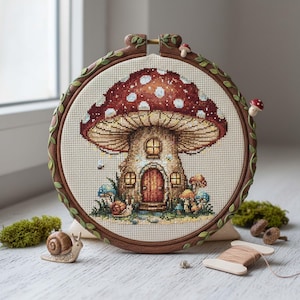 Könnte beinhalten: Kreuzstich-Stickerei eines Pilzhauses mit rotem und weiß gepunktetem Hut, braunem Stiel und Holztür. Die Stickerei befindet sich in einem Holzreifen mit dekorativem Rankenrand. Zusätzliche Pilze und eine Schneckenfigur befinden sich in der Nähe.