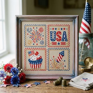 May include: Framed cross-stitch artwork featuring patriotic imagery. The design includes fireworks, the word "USA," a cupcake with a flag, and a rocket, all in red, white, and blue. The artwork is displayed on a wooden surface with additional patriotic decorations.