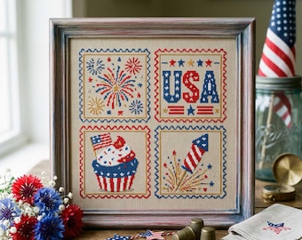 May include: Framed cross-stitch artwork featuring patriotic imagery. The design includes fireworks, the word "USA," a cupcake with a flag, and a rocket, all in red, white, and blue. The artwork is displayed on a wooden surface with additional patriotic decorations.