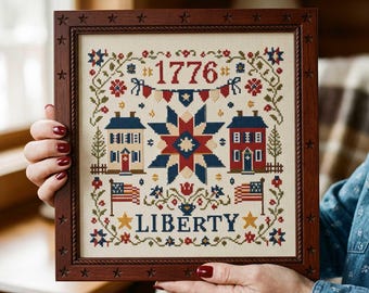 May include: Cross-stitch artwork in a dark wooden frame with star accents. The design features a central star, two houses, flags, and the word "LIBERTY" below the year "1776". The color palette includes red, white, blue, and yellow.