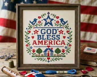 May include: A framed cross-stitch piece with the text "GOD BLESS AMERICA" in red and blue. The design includes a star, birds, flowers, and a house, set against an American flag background. Craft supplies are in the foreground.