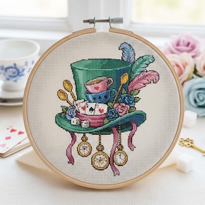 May include: A cross-stitch embroidery in a wooden hoop depicts a whimsical top hat design. The hat is teal with pink ribbons, decorated with teacups, playing cards, pocket watches, and feathers. The colour palette includes teal, pink, blue, and gold.