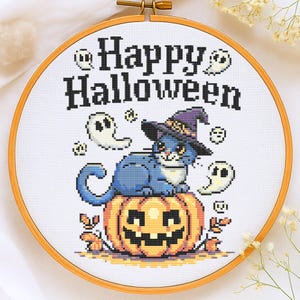 Pode incluir: Um bastidor de bordado em ponto cruz com tema de Halloween. Apresenta um gato azul com um chapéu de bruxa em cima de uma abóbora, com as palavras "Happy Halloween" e motivos de fantasmas e folhas.