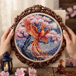 May include: A cross-stitch embroidery of a phoenix with outstretched wings, set against a backdrop of cherry blossoms and a full moon. The artwork is framed in a decorative, ornate, brown wooden frame. The phoenix is rendered in shades of orange and red.