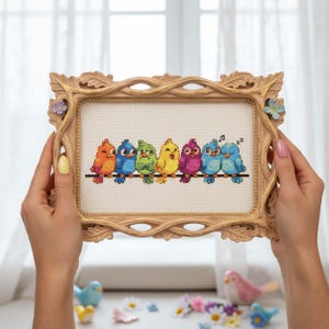 Finished Rainbow Birds Cross Stitch Pattern displayed in a hoop shows seven bright birds perched on a wire, each stitched in a different rainbow shade. Their playful personalities include singing, winking, and dozing off, making the design full of charm. The vibrant palette features oranges, yellows, blues, greens, and purples, balanced with backstitch outlines for clarity. This counted embroidery PDF pattern is perfect for stitchers who love cheerful and whimsical art, creating a handmade