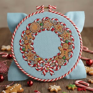 May include: A cross-stitch embroidery hoop with a festive Christmas wreath design. The wreath includes gingerbread men, star-shaped biscuits, and candy canes, set against a light blue fabric. The hoop is trimmed with red and white striped fabric.