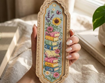 May include: A wooden bookmark with a cross-stitched design featuring a stack of colourful books, flowers, and a sunflower. The bookmark has a light blue background and is framed with a decorative wooden border.