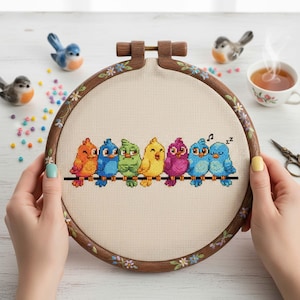 Puede incluir: Un bastidor de bordado de punto de cruz con siete pájaros coloridos posados ​​en una línea negra. Los pájaros son en tonos de naranja, azul, verde, amarillo y morado. El aro de madera está decorado con flores pintadas. El fondo es una tela beige claro.