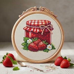 Op de afbeelding: Kruissteekborduurwerk van een pot aardbeienjam met een rood-wit geblokte deksel en een strik. De borduursel zit in een houten hoepel, met verse aardbeien en een lepel in de buurt. De jam pot is omgeven door groene bladeren en witte bloemen.