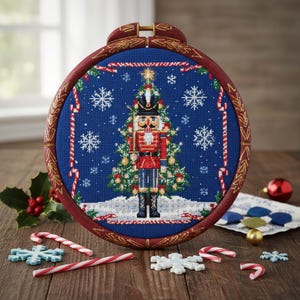 Puede incluir: Un bastidor de bordado de punto de cruz con un diseño navideño. Muestra un cascanueces y un árbol de Navidad sobre un fondo azul. El bastidor de madera es rojo oscuro con detalles dorados. Bastones de caramelo y copos de nieve decoran la escena.