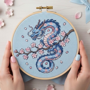 May include: Cross-stitch embroidery of a blue dragon with pink accents and cherry blossoms on a light blue background, set in a wooden hoop. The dragon is wrapped around a flowering branch. The hands are holding the embroidery hoop.