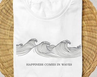 Könnte beinhalten: Weißes T-Shirt mit einer schwarzen Strichzeichnung von Wellen. Unter den Wellen steht der Text "HAPPINESS COMES IN WAVES". Das T-Shirt liegt gefaltet auf einer runden Matte aus Naturfasern.