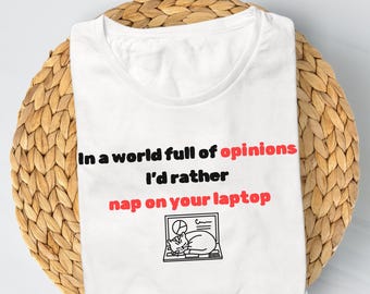Op de afbeelding: Wit T-shirt met de tekst "In a world full of opinions I'd rather nap on your laptop" in zwart en rood. Een kleine afbeelding van een kat die op een laptop dut, staat eronder. Het shirt ligt op een geweven, ronde placemat.