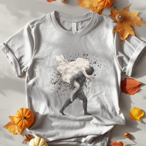 Puede incluir: Una camiseta gris claro con una ilustración en escala de grises de una persona que lleva a otra persona. El diseño tiene un efecto de salpicadura. La camiseta está rodeada de hojas de otoño y pequeñas calabazas.