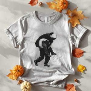 Puede incluir: Una camiseta gris claro con un gráfico en blanco y negro de una persona cargando a otra sobre su espalda. La camiseta está rodeada de hojas de otoño y pequeñas calabazas.