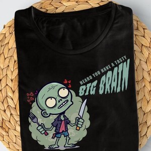 Könnte beinhalten: Schwarzes T-Shirt mit einem Cartoon-Zombie, der eine Gabel und ein Messer hält, mit dem Text "HEARD YOU HAVE A TASTY BIG BRAIN." Das T-Shirt wird auf einer gewebten Matte präsentiert.