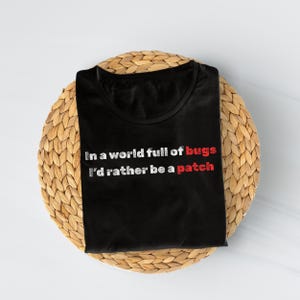 May include: Black t-shirt with the text "In a world full of bugs I'd rather be a patch" in white and red lettering. The shirt is folded and placed on a round, woven, light brown placemat.
