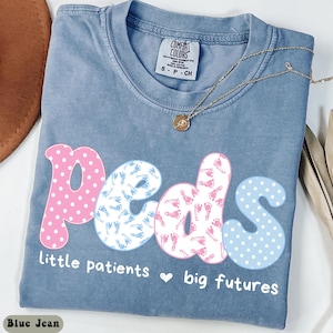 May include: Blue jean colored t-shirt with the word "Peds" in pink and blue lettering. The letters are decorated with polka dots and baby footprints. The text "Little patients, big futures" is below the word "Peds". A gold necklace is draped across the shirt.