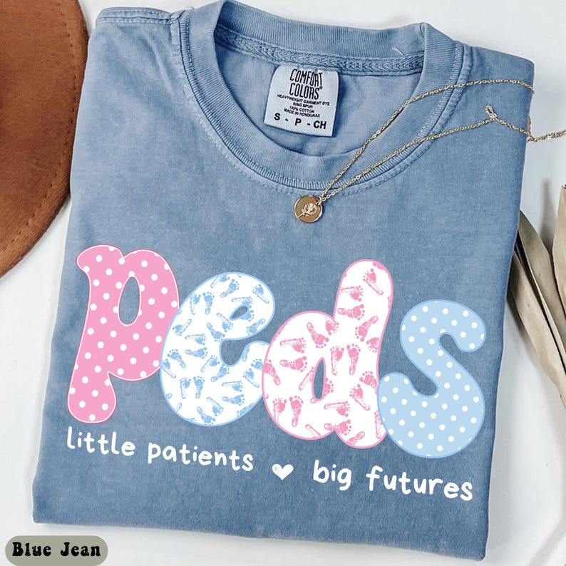 May include: Blue jean colored t-shirt with the word "Peds" in pink and blue lettering. The letters are decorated with polka dots and baby footprints. The text "Little patients, big futures" is below the word "Peds". A gold necklace is draped across the shirt.
