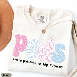 May include: Ivory-colored t-shirt with the word "peds" in pink and blue, featuring polka dots and baby footprints. The text "little patients ❤️ big futures" is below the word "peds". A gold necklace is draped across the shirt.