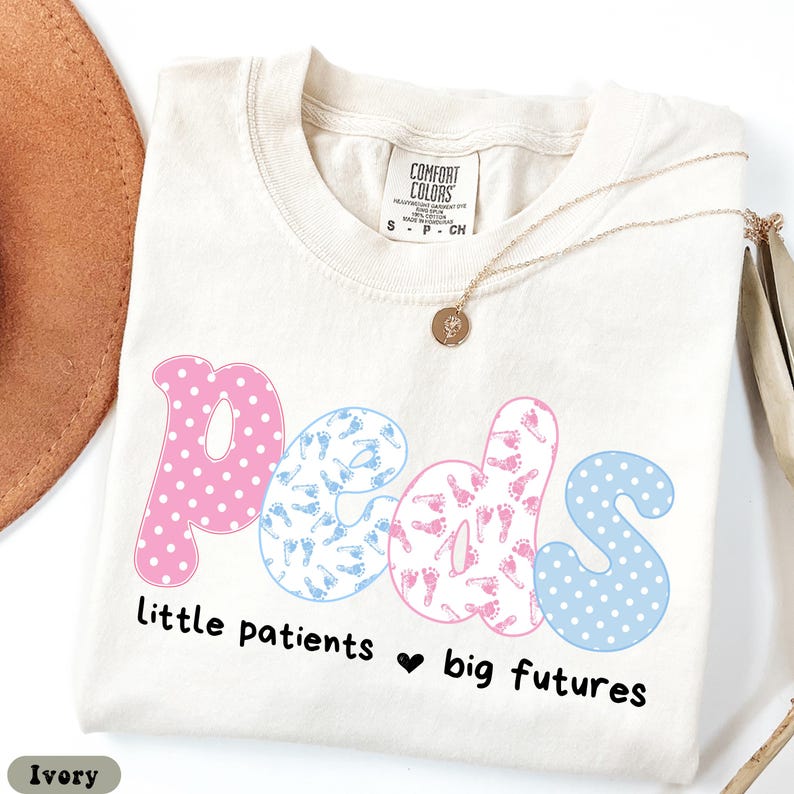May include: Ivory-colored t-shirt with the word "peds" in pink and blue, featuring polka dots and baby footprints. The text "little patients ❤️ big futures" is below the word "peds". A gold necklace is draped across the shirt.