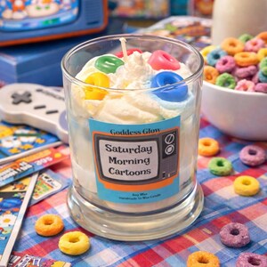 Op de afbeelding: Een glazen kaars met een witte wasbasis, versierd met slagwas en kleurrijke, graanvormige wasaccenten. De kaars heeft een label met de tekst "Saturday Morning Cartoons". Andere items zijn een kom met ontbijtgranen, een retro tv en stripboeken.