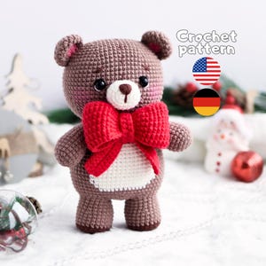 May include: A brown crocheted teddy bear with a white muzzle, black eyes, and a large red bow around its neck. The bear has a white belly patch and pink blush on its cheeks. The text "Crochet pattern" is visible with US and German flag icons.