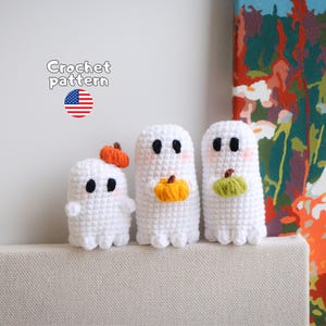 May include: Three white crocheted ghost figures, each with black eyes and pink cheeks. The smallest ghost wears an orange pumpkin on its head, while the two larger ghosts hold small orange and green pumpkins. Text reads Crochet pattern.