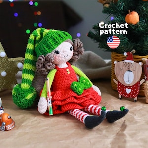 Puede incluir: Una muñeca de ganchillo con pelo rizado castaño, que lleva un gorro largo a rayas verdes y lima con un pompón, un vestido rojo con botones y lazo verdes, y mallas a rayas rojas y blancas con zapatos negros. El texto Crochet pattern es visible.
