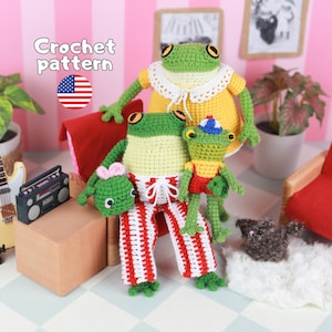 May include: Three crocheted frog dolls. The largest wears a yellow dress with a white lace collar. The middle frog wears red and white striped pants with a white drawstring. The smallest frog wears a yellow shirt, red overalls, and a blue hat.