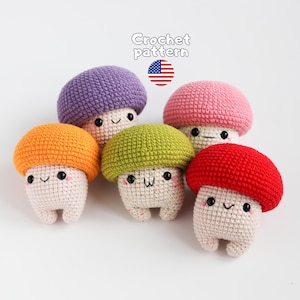 May include: Five small crochet mushroom plushies with cream-colored stems and caps in purple, pink, orange, green, and red. Each has black bead eyes, a stitched smile, and pink cheeks. Text above reads Crochet pattern with a US flag icon.