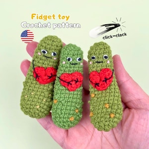 Op de afbeelding: Drie gehaakte, augurkvormige speeltjes in het groen, elk voorzien van een rood hartje, geborduurde oogjes en kleine gele vlekjes. De afbeelding bevat de tekst Fidget toy Crochet pattern en click-clack naast een zwart-wit haarspeldicoon.