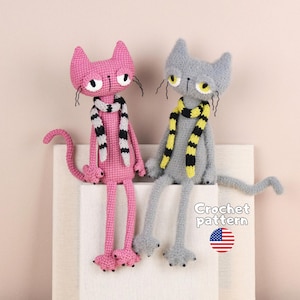 Könnte beinhalten: Zwei handgefertigte Häkelkatzen, eine rosa und eine grau, beide mit gestreiftem Schal. Die rosa Katze trägt einen schwarz-weißen Schal, die graue einen gelb-schwarzen. Text Crochet pattern mit einem kleinen US-Flaggen-Symbol.