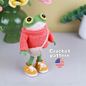 May include: A crocheted frog figurine with green skin, large yellow and black eyes, wearing a coral-colored sweater, a small beige crossbody bag with a button, and yellow sneakers with white laces. Text reads Crochet pattern with a US flag icon.