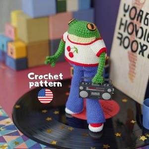 May include: A crocheted frog figurine wearing a white t-shirt with a red cherry graphic and blue pants, holding a miniature black boombox. The text Crochet pattern is visible next to a small United States flag icon on the left side of the image.
