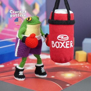May include: A green and yellow crocheted frog wearing purple boxing shorts and black boots with white laces. The frog has red crocheted boxing gloves and is positioned next to a red hanging punching bag with the word BOXER embroidered in white.
