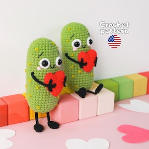 Könnte beinhalten: Zwei grüne, gehäkelte Gurkenfiguren mit schwarzen Augen, rosa Wangen und kleinen schwarzen Gliedmaßen. Jede Figur hält ein leuchtend rotes, gehäkeltes Herz. Der Text "Crochet pattern" und eine kleine US-Flagge sind oben rechts zu sehen.
