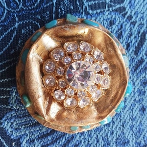 Könnte beinhalten: Eine goldfarbene Brosche mit einem großen, klaren Strassstein, der von kleineren Strasssteinen umgeben ist. Die Brosche ist mit türkisfarbenen Steinen am Rand verziert.