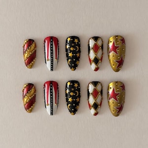 May include: A set of ten almond-shaped press-on nails featuring diverse designs including red and gold diagonal stripes, red and white vertical panels with studs, black with gold stars and moons, white and red harlequin patterns, and gold stars.