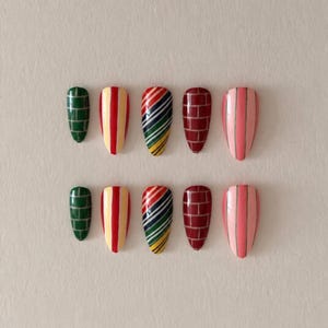 Può includere: Un set di dieci unghie finte a forma di mandorla disposte su due file. I design includono verde scuro con motivo a mattoni bianco, strisce verticali rosse e gialle, strisce diagonali multicolori, rosso scuro con motivo a mattoni bianco e rosa con strisce verticali.