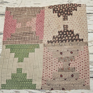 May include: A patchwork quilt square with four distinct sections. Each section features a log cabin design with various patterned fabrics in shades of brown, pink, and green. The quilt is placed on a white wooden surface.