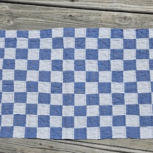 May include: A rectangular quilted fabric piece featuring a repeating checkerboard pattern of alternating blue and white squares. The textile has visible stitching lines that define each square, creating a textured, soft, and layered appearance.
