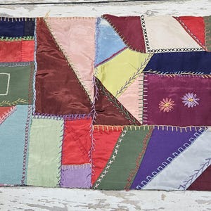 May include: A colorful patchwork textile with a variety of fabric pieces in shades of red, blue, green, purple, and pink. The pieces are sewn together with visible stitching, and some have embroidered details. The textile is likely a quilt or decorative piece.