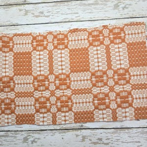 May include: A rectangular woven textile mat featuring a repeating geometric pattern in burnt orange and off-white. The fabric displays a dense, textured weave with alternating blocks of color that create a structured, grid-like visual composition.