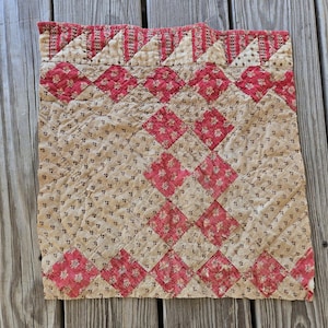 May include: A square quilted textile featuring a repeating diamond pattern in muted red and beige floral prints. The top border displays a row of alternating red and beige triangles, with visible stitching throughout the fabric surface.